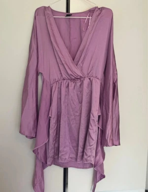 Ljuslila omlottklänning med volang - Säljer en ljuslila omlottklänning med v-ringning och långa ärmar. Klänningen har volangdetaljer och asymmetrisk nederkant som ger en flowy känsla. Perfekt för dig som gillar en feminin och trendig stil.