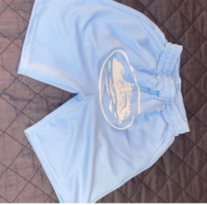 Ljusblåa loose shorts med tryck - Säljer ett par ljusblåa shorts med bred resår i midjan och ett vitt tryck framtill. Modellen är loose fit och har en bakficka. Perfekta för sommaren och chill dagar.