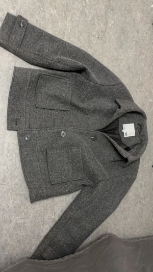 Grå overshirt från 157, storlek 160 - Snygg grå overshirt från 157 med klassisk krage och stora fickor framtill. Jackan har knappar och är helfodrad med svart tyg på insidan. Perfekt för dig som gillar stilrena och enkla plagg med lite edge.