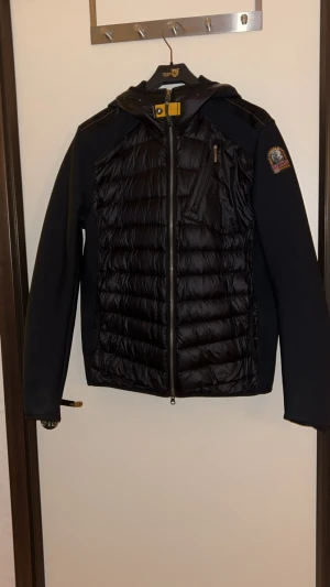 Svart pufferjacka Parajumpers M - Svart pufferjacka från Parajumpers med huva och coolt quiltat mönster på både fram- och baksida. Jackan har dragkedja, bröstficka med zip och patch på ärmen. Insidan har orange detaljer och flera fickor. Perfekt för dig som vill ha en snygg och funktionell jacka.