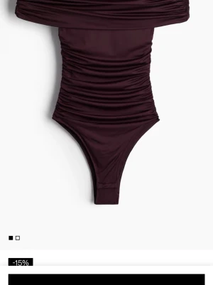 Mörklila offshoulder bodysuit - Säljer en mörklila bodysuit med offshoulder-design och snygga rynkade detaljer längs sidorna. Materialet är stretchigt och följsamt, vilket ger en tight passform. Perfekt att styla med höga jeans eller kjol.