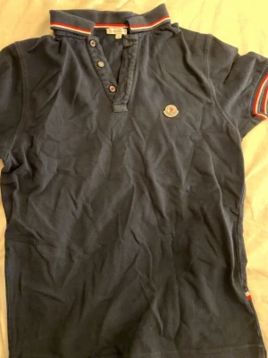 Mörkblå Moncler pikétröja S - Säljer en mörkblå pikétröja från Moncler i storlek S. Tröjan har klassisk krage med röd och vit randig detalj, korta ärmar och knappar framtill. Moncler-logga på bröstet och mjukt bomullsmaterial. Perfekt till en avslappnad men ändå snygg look.