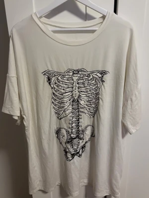 Vit oversized t-shirt med skelettprint - Säljer en vit oversized t-shirt med ett stort svart skelettmotiv på framsidan. T-shirten har korta ärmar och rund halsringning. Storlek står inte men skulle säga L