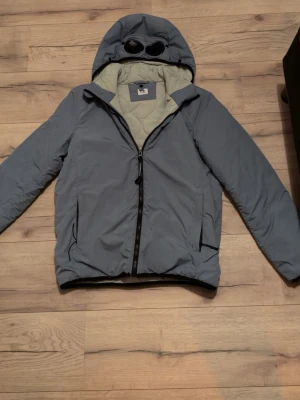 Blå C.P. Company pufferjacka med goggles - Säljer en blå pufferjacka från C.P. Company med deras ikoniska goggles i huvan. Jackan har quiltad insida, dragkedja framtill och två sidofickor. Tillverkad i syntetmaterial och perfekt för kyligare dagar. Stilsäker och funktionell jacka för dig som gillar streetwear.