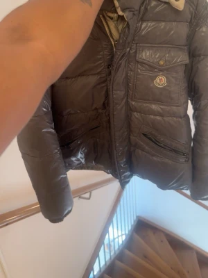 Mörkgrå Moncler pufferjacka - Säljer en mörkgrå pufferjacka från Moncler med blank finish och flera fickor med dragkedja. Jackan har huva, långa ärmar och klassisk Moncler-logga på bröstet. Perfekt för kalla dagar och riktigt snygg till streetwear-stilen. Går att vända ut och in ifall man vill använda den beiga insidan även! Helt ny skick använd fåtal gånger! Pris kan diskuteras vid snabb affär, 4500kr. Size 3 