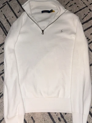Vit half zip tröja från Polo Ralph Lauren - Vit långärmad tröja från Polo Ralph Lauren med half zip och diskret broderad logga på bröstet. Tröjan är i mjuk bomull och har en klassisk, stilren look som passar perfekt till jeans eller chinos.