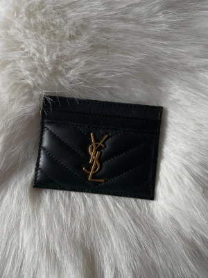 Svart quiltad plånbok i skinn - Säljer en svart quiltad plånbok med ikonisk guldlogga på framsidan. Plånboken är tillverkad i skinn och har flera fack för kort. Snygg och stilren design som passar perfekt i fickan eller väskan.