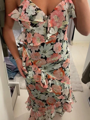 Blommig långklänning med volanger - Säljer en superfin långklänning med stora blommor i rosa, vit och grönt på svart botten. Klänningen har volangdetaljer vid axlar och längs kjolen, v-ringad hals och smala axelband. Materialet känns lätt och luftigt, perfekt för varma dagar.