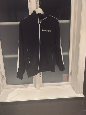 Palm Angels svart zip hoodie - Svart hoodie från Palm Angels med vit dragkedja och vita ränder längs ärmarna. Tröjan har hög krage, två fickor med dragkedja och logotyp på bröstet. Perfekt för dig som gillar streetwear och vill ha en snygg och bekväm look.