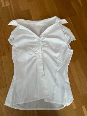 Vit kortärmad skjorta med knappar - Snygg vit skjorta med korta ärmar och klassisk krage. Skjortan har knappar framtill och fina rynkade detaljer vid bysten som ger en schysst form. Perfekt till jeans eller kjol för en clean look.