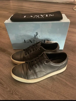 Lanvin Sneakers – Svart Läder, Herr – Klassisk Modell - Säljer ett par Lanvin herrsneakers i svart läder med vit sula. En äldre, klassisk modell med riktigt bra kvalitet och stilren design. Skorna är använda men i fint skick för sin ålder – se bilder.  Info  • Märke: Lanvin • Färg: Svart med vit sula • Material: Äkta läder • Storlek: 43 • Skick: Använda, men välvårdade