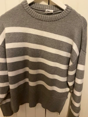 Gråvit randig stickad tröja  - Säljer en stickad tröja i grått och vitt med breda horisontella ränder. Tröjan har rund halsringning, ribbade muddar och är långärmad. Perfekt för kyliga dagar och enkel att matcha med jeans eller kjol.