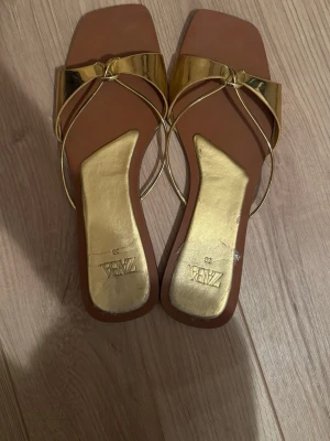 Guldiga sandaler från Zara - Säljer ett par sandaler från Zara i metallic guld med fyrkantig tå och tunna remmar. Snygg design med platt sula och glansigt material som verkligen sticker ut. Perfekta för sommardagar när du vill ha något extra.