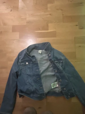 Blå jeansjacka från &Denim - Snygg blå jeansjacka från &Denim med klassisk krage, knappar framtill och bröstfickor med lock. Jackan har långa ärmar och är tillverkad i slitstarkt denimtyg. Perfekt för dig som vill ha en tidlös och cool look.