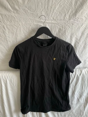 Svart t-shirt från Lyle & Scott - Klassisk svart t-shirt från Lyle & Scott med gul broderad logga på bröstet. T-shirten har rund halsringning och korta ärmar. Tillverkad i mjuk bomull för en skön känsla och enkel stil.