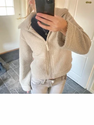 Beige teddyjacka med dragkedja - Supermjuk och mysig teddyjacka i beige med hel dragkedja framtill och två stora fickor. Jackan har hög krage och långärmad passform, perfekt för kyliga dagar. Materialet är fluffigt och ger en chill vibe till din outfit.