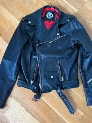 Svart skinnjacka med bälte och dragkedjor - Säljer en svart äkta skinnjacka från Shock med röd insida, klassisk bikerstil och flera dragkedjor. Jackan har bälte i midjan, snedställd dragkedja framtill och fickor med dragkedjor. Snygga detaljer som axelklaffar och dragkedjor vid ärmslut. Är lite sliten på vänstra ärmen. Se bild.