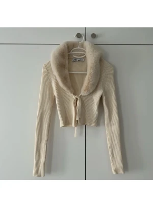 Beige croppad kofta med fuskpäls från Zara - Säljer en beige croppad kofta från Zara med ribbad struktur och stor mjuk fuskpäls-krage. Koftan har långa ärmar och knytband framtill för en extra söt detalj. Perfekt för dig som vill ha en trendig och cozy look.