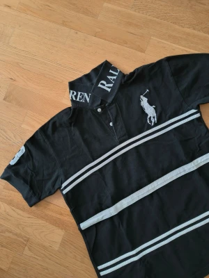 Svart polotopp från Ralph Lauren - Säljer en svart polotopp från Ralph Lauren med vita ränder och stor logga på bröstet. Kragen har tydlig text med märkesnamnet och toppen har korta ärmar samt knappar framtill. Tillverkad i  bomull, perfekt för en sportig och stilren look.