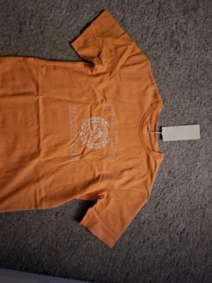 Orange t-shirt Move Studio - Snygg orange t-shirt från Move Studio med tryck på bröstet där det står 'Racquet Club'. Klassisk rund hals och korta ärmar. Tillverkad i mjuk bomull som känns skön mot huden. Perfekt för dig som gillar sportig och avslappnad stil.