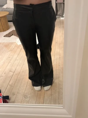 Svarta bootcut byxor i skinnimitation - Säljer ett par svarta byxor i skinnimitation med bootcut passform och hög midja. Byxorna har en clean look utan synliga detaljer och sitter snyggt över höfterna. Perfekta för dig som vill ha en edgy vibe till din outfit.