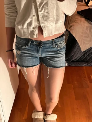 Blå jeansshorts från Abercrombie - Säljer ett par blå jeansshorts från Abercrombie med låg midja. De har slitna detaljer, fransig rå kant och klassisk femficksdesign. Perfekta för sommaren och ger en avslappnad vibe. Materialet är jeans och shortsen har knapp- och dragkedjestängning. Jag har storlek 25 i midjan och dom är lite för små och sitter rätt tight.