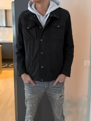 Svart jacka från Peak Performance L - Säljer en svart jacka från Peak Performance i storlek L. Jackan har flera fickor framtill med tryckknappar och klassisk krage. Materialet är polyester och insidan är quiltad för extra komfort. Perfekt för dig som gillar stilren och funktionell design.