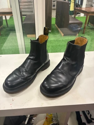 Svarta Chelsea boots från Dr. Martens - Svarta Chelsea boots i skinn från Dr. Martens med klassisk rund tå och resår på sidorna. Snygg gul logotyp på flärpen bak och kontrastfärgad insida. Perfekta för dig som vill ha en stilren och tidlös look.