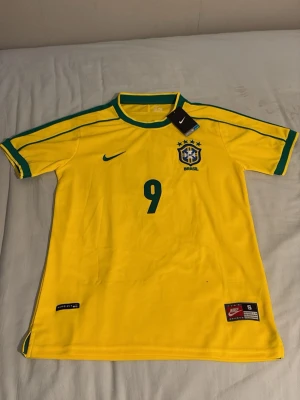 Brasilien Ronaldo 9 Nike matchtröja - Gul Brasilien 1998 matchtröja från Nike med gröna detaljer, nummer 9 och Ronaldo på ryggen. Aldrig använd eftersom den är för stor för mig. Klassisk design med rund hals och broderad landslagssköld. Perfekt för dig som älskar fotboll och vill hylla en ikon. 