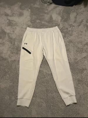 Vita mjukisbyxor från Under Armour XL - Säljer ett par vita mjukisbyxor från Under Armour i storlek XL. Byxorna har resår i midjan, ribbade muddar vid bensluten och en svart dragkedja på ena fickan. Perfekta för chill eller träning, riktigt sköna och stilrena.