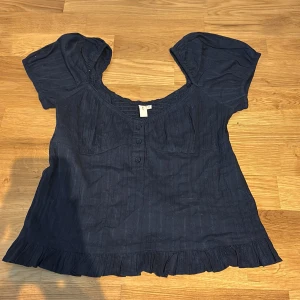 Mörkblå blus med volang från Nelly - Supersöt mörkblå blus från Nelly med kort puffärm, knappar framtill och volangkant nedtill. Blusen har en lätt rynkad struktur och smockdetalj vid halsringningen. Perfekt för dig som gillar romantiska detaljer och en luftig känsla.