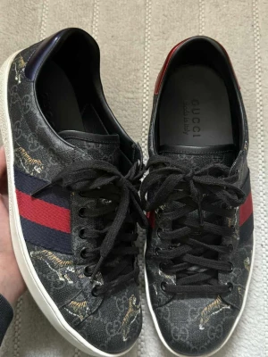 Gucci sneakers - Snygga svarta Gucci sneakers med klassiskt rött och marinblått band på sidan. Skorna har ett coolt tigerprint och GG-mönster över hela ovandelen. Tillverkade i skinn och textil med svarta snören och vit sula. Perfekta för dig som vill sticka ut med lyxig streetstyle. Storlek 8 , passar 42,5-43