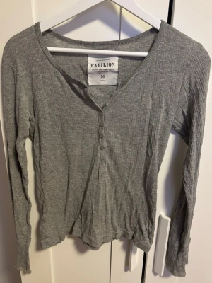 Grå ribbad långärmad topp Fabilion - Grå långärmad topp från Fabilion. Modellen har v-ringning med knappar framtill och en liten broderad detalj på bröstet. Mjuk och stretchig passform som sitter snyggt på kroppen. 