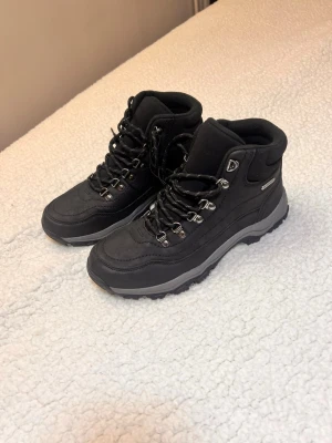 Svarta vattentäta kängor med snörning - Svarta kängor med grov sula och snörning, perfekta för utomhusaktiviteter. Skorna är vattentäta och har detaljer i metall vid snörningen. Ovandelen är i syntetmaterial och insidan är vadderad för extra komfort. Snygg grå kontrast på sulan.