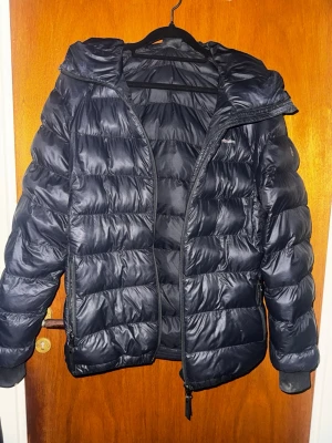 Svart pufferjacka Peak Performance - Säljer en svart pufferjacka från Peak Performance med huva och dragkedja. Jackan har en quiltad design och är tillverkad i syntetmaterial. Perfekt för kalla dagar och riktigt skön att bära.