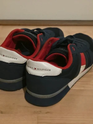 Tommy Hilfiger sneakers i marinblått - Snygga sneakers från Tommy Hilfiger i marinblått med röda och vita detaljer. Skorna har röd insida, vita och röda paneler på sidan samt klassisk snörning. Ovandelen är i en mix av mocka och mesh för en sportig look.