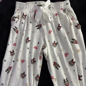 Vita julmönstrade mjukisbyxor  - Supermysiga vita mjukisbyxor med gulligt julmönster av renar, stjärnor och snöflingor. Byxorna har resår i midjan, bred knytrosett och muddar vid bensluten. Perfekta för chilliga vinterkvällar och julmys!