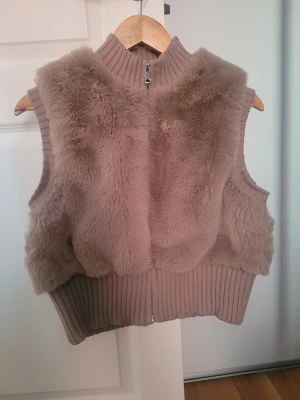 Beige pälsväst från Bershka - Säljer en beige pälsväst från Bershka i storlek L. Västen har mjuk fuskpäls på utsidan, ribbade kanter och hög krage. Stängs med dragkedja framtill och har en croppad passform som ger en trendig look.