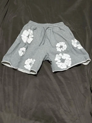 Grå blommiga shorts från Denim Tears - Grå shorts från Denim Tears med vita blommönster över hela plagget. De har elastisk midja med snörning och är tillverkade i mjuk bomull, perfekta för chill dagar. Passformen är loose och de har en avslappnad vibe.