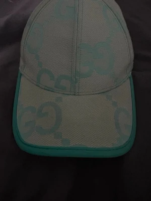 Mintgrön keps med GG-mönster - Säljer en snygg mintgrön keps med stort GG-mönster över hela kepsen. Kepsen har justerbar rem bak och är tillverkad i bomull och polyester. Perfekt accessoar för att lyfta din outfit med en lyxig touch.