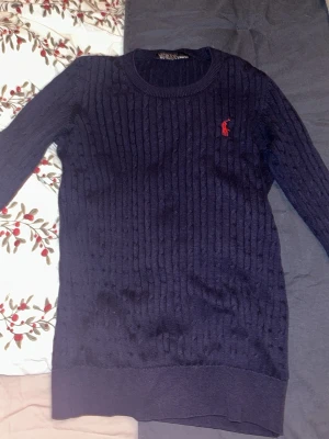 Mörkblå kabelstickad tröja Polo Ralph Lauren - Kabelstickad mörkblå tröja från Polo Ralph Lauren med klassisk röd logga broderad på bröstet. Tröjan har rund halsringning, lång ärm och ribbade muddar. Perfekt till höst och vinter för en stilren look.  PRIS KAN DISKUTERAS!