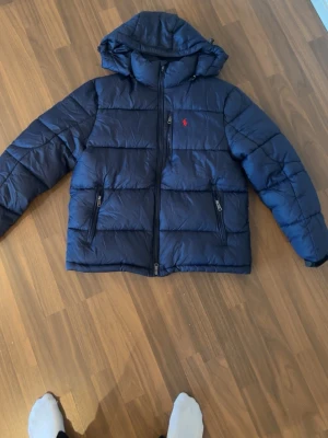 Mörkblå pufferjacka från Polo Ralph Lauren - Säljer en mörkblå pufferjacka från Polo Ralph Lauren med huva och två dragkedjeförsedda fickor. Jackan har det klassiska röda Polo-logot broderat på bröstet och grov dragkedja framtill. Perfekt för kalla dagar och riktigt snygg till streetwear.