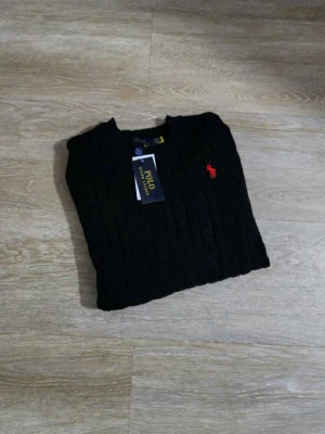 Svart kabelstickad tröja Polo Ralph Lauren - Kabelstickad svart tröja från Polo Ralph Lauren med klassisk röd logga broderad på bröstet. Tröjan har rund halsringning och långa ärmar. Perfekt till höst och vinter för en clean och stilren look.