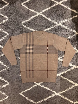 Beige rutig stickad tröja - Säljer en beige stickad tröja med rutigt mönster i bruna och grå toner. Tröjan har rund halsringning, ribbade muddar och lång ärm. Jag väger 70KG och är 180CM och den är stor på mig. 