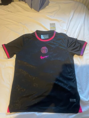 Svart PSG x Nike fotbollströja S - Snygg svart Paris Saint-Germain fotbollströja från Nike med rosa/lila detaljer och logga. Tröjan har korta ärmar, rund hals med rosa kant och subtilt blommönster i tyget. Materialet är lätt och andas, perfekt för träning eller match.