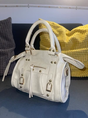 Vit handväska med gulddetaljer - Snygg vit handväska med guldfärgade nitar och spännen. Väskan har dubbla handtag, dragkedja upptill och en liten ficka med dragkedja framtill. Insidan är ljusbeige och har en innerficka. Kommer med tillhörande axelband som fästs på sidan av väskan Perfekt storlek för det viktigaste och ger en clean look. Köpt i Italien. Aldrig använd. Säljer pga av att det inte är min stil💞