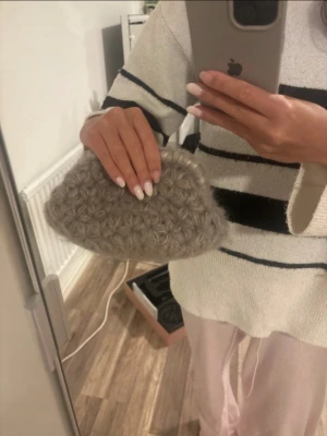 Grå virkad clutchväska - Säljer en superfin grå clutchväska i fluffigt virkat material. Väskan har ett unikt virkat mönster och stängs med små guldfärgade detaljer upptill. Perfekt storlek för det viktigaste och ger en mysig touch till din outfit.