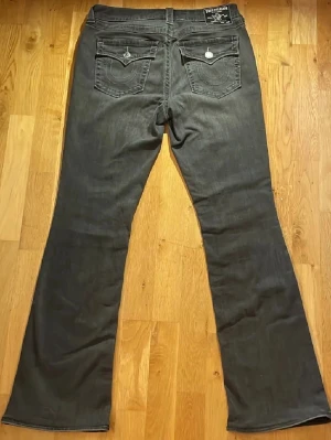 True Religion grå bootcut jeans - Säljer ett par gråa bootcut jeans från True Religion, modell Becca. Jeansen har klassiska kontrastsömmar, fem fickor och snygga detaljerade bakfickor med lock och silverknappar. Tillverkade i mjuk bomullsdenim med en riktigt schysst passform. Storlek: W30/L-Xl. Modell: Becca