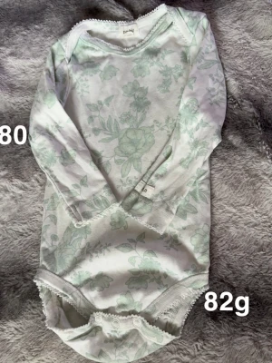 Blommig långärmad body Newbie - Säljer en söt långärmad body från Newbie i storlek 80. Bodyn är vit med ljusgröna blommor och har fina spetsdetaljer vid hals och ben. har tryckknappar i grenen för enkel påklädning.🌸 Fint använt skick. Storlek 80