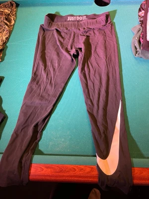Svarta Nike leggings med stor logga - Svarta leggings från Nike i storlek L med stor vit swoosh-logga längs ena benet och texten 'JUST DO IT.' i midjan. Tight passform och tillverkade i stretchigt syntetmaterial, perfekta för träning eller chill. Klassisk sportig look.